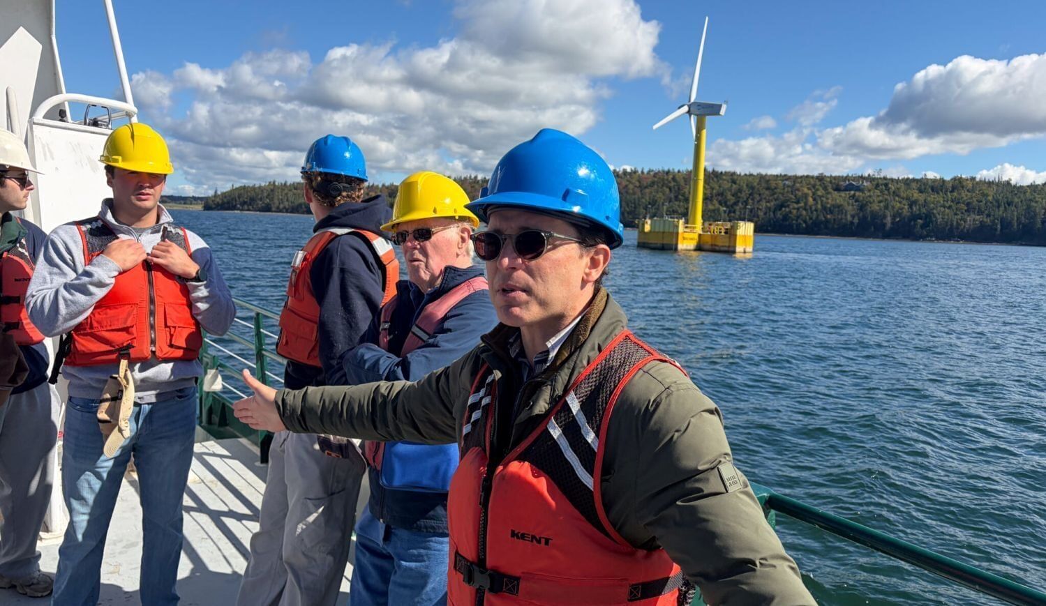 People in hard hats stand on a boat with a wind turbine behind them