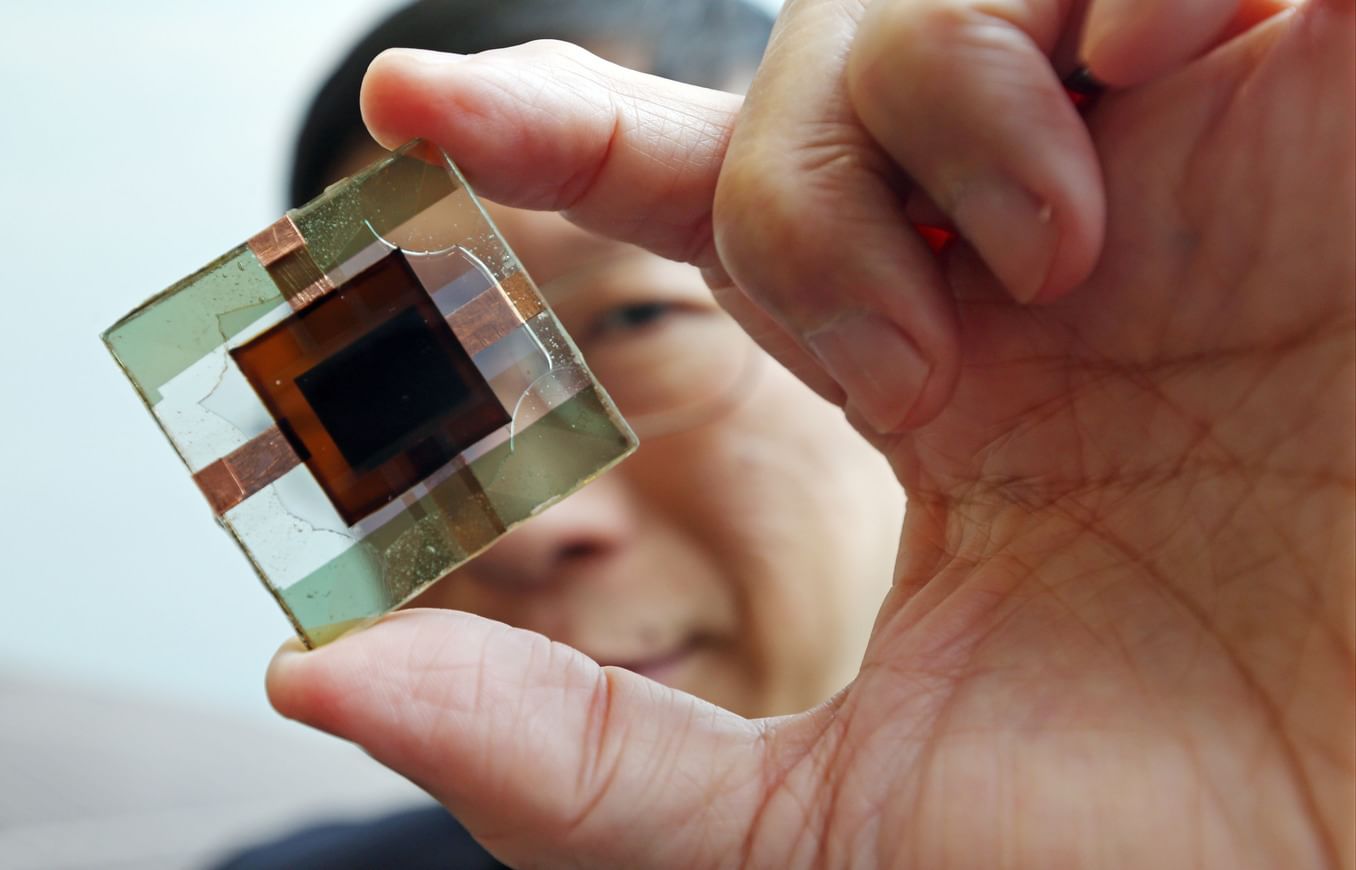 A man with black hair and wire-rimmed glasses holds a small glass square with a smaller square of dark brown material inside