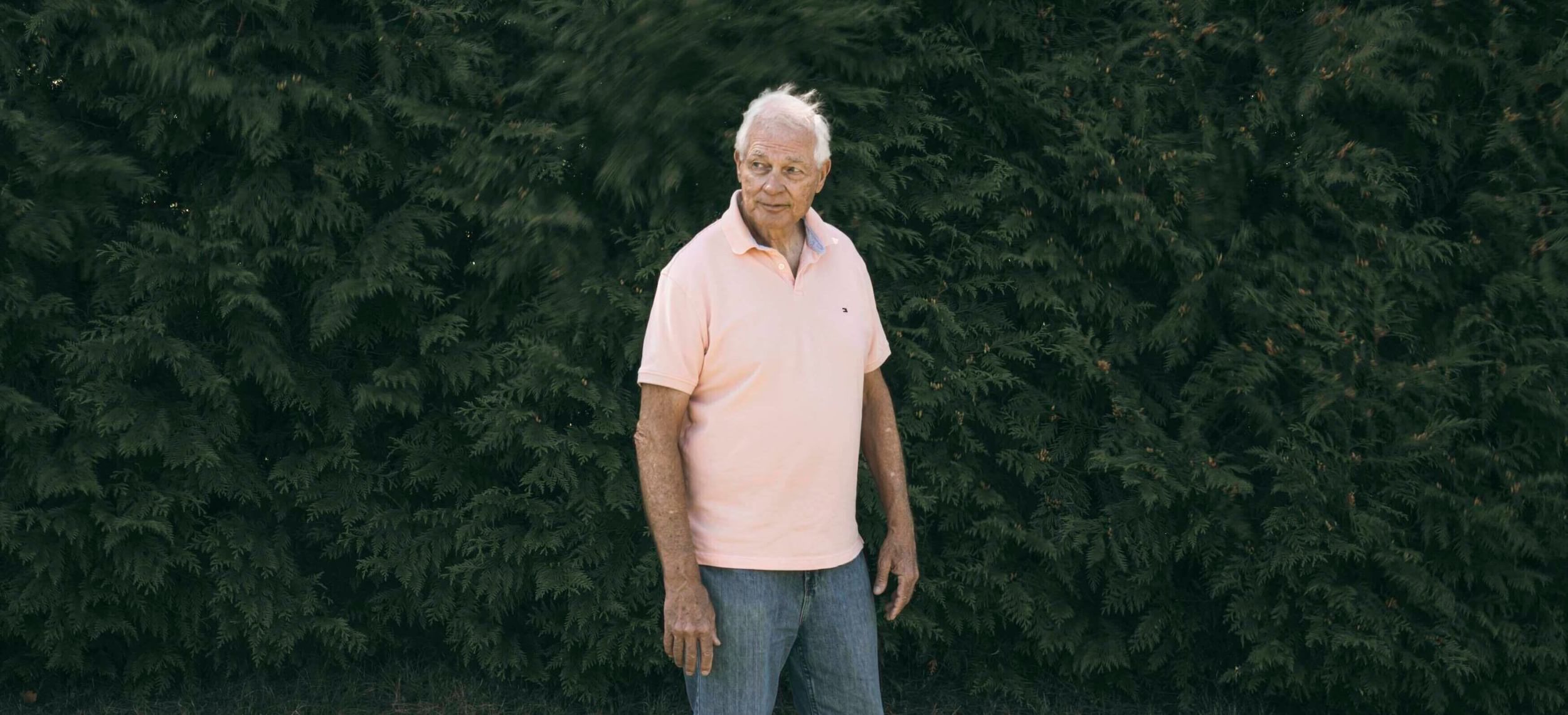 A man in a light-pink shirt stands before a wall of greenery.